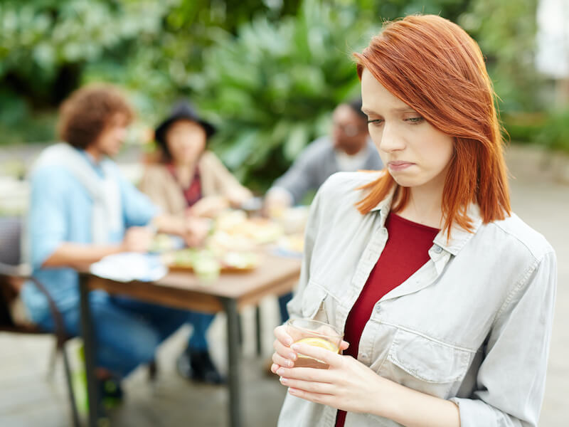 Unhappy girl suffering from hearing loss with glass of drink expressing sadness and loneliness while her friends having fun and enjoying a festive BBQ dinner in the background