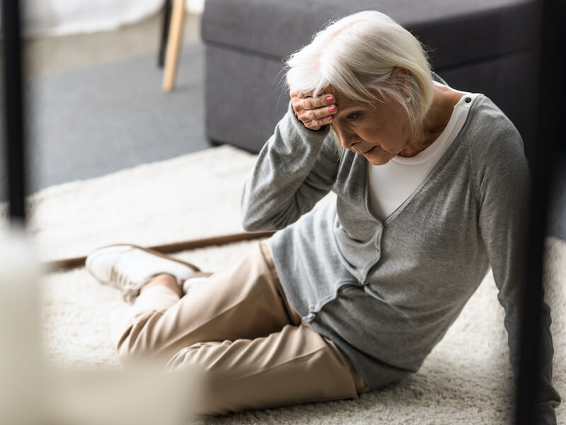 Senior woman fell down and is sitting on carpet and touching forehead with hand 