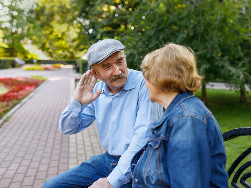 Elderly man leans in and cups ear to try to hear his spouse while sitting on a park bench